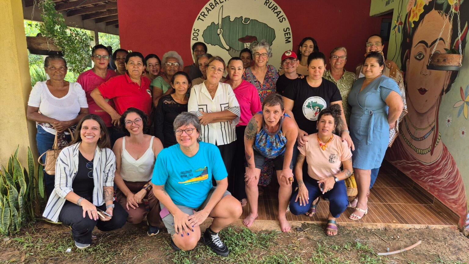 Cerca de trinta mulheres reunidas e sorrindo diante de um mural com o rosto de Frida Kahlo, num espaço do Movimento dos Trabalhadores Rurais Sem Terra